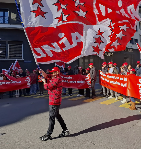 Une manifestation d'Unia à Sion avec en tête de cortège un gigantesque drapeau composé du logo d'Unia et des armes du canton du Valais, tenu par un membre d'Unia