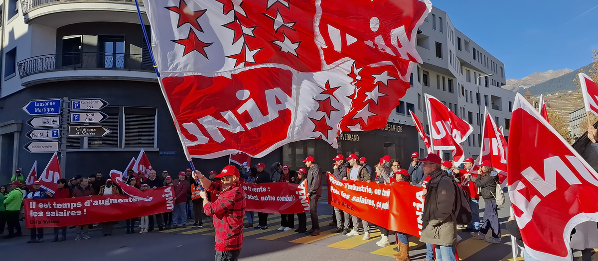Une manifestation d'Unia à Sion avec en tête de cortège un gigantesque drapeau composé du logo d'Unia et des armes du canton du Valais, tenu par un membre d'Unia